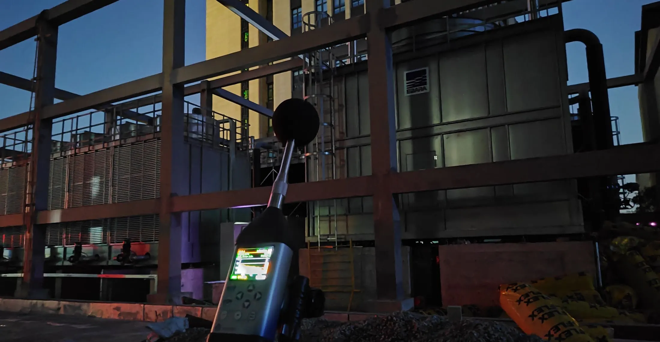 Sound level meter measuring cooling tower noise on a construction site before dawn